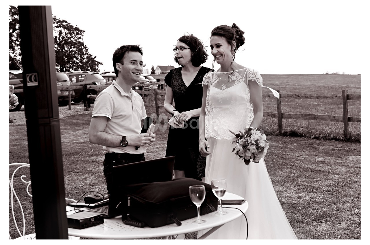 Un homme avec un micro discute avec une mariée souriante et une femme, en extérieur près d'une table équipée.