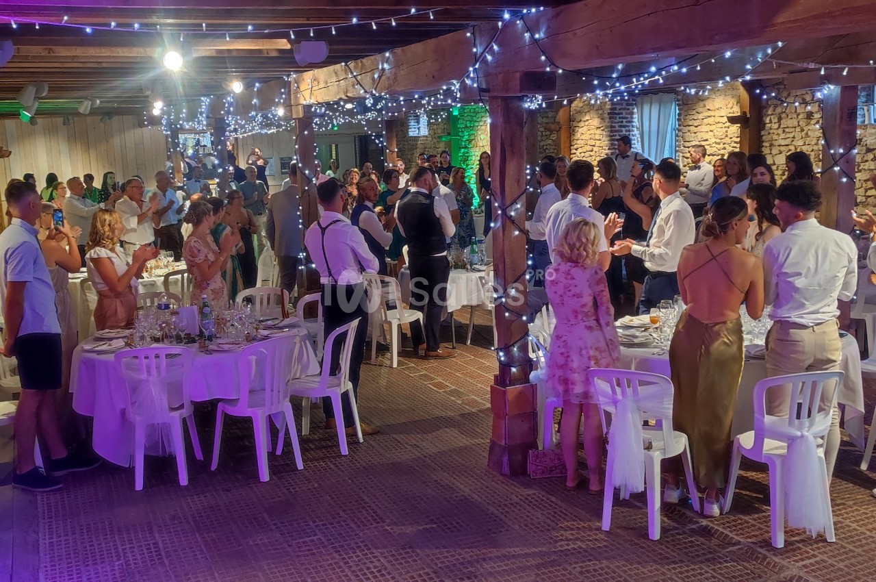 Salle de réception avec des invités debout et assis, décorée de guirlandes lumineuses et murs en pierre apparente.