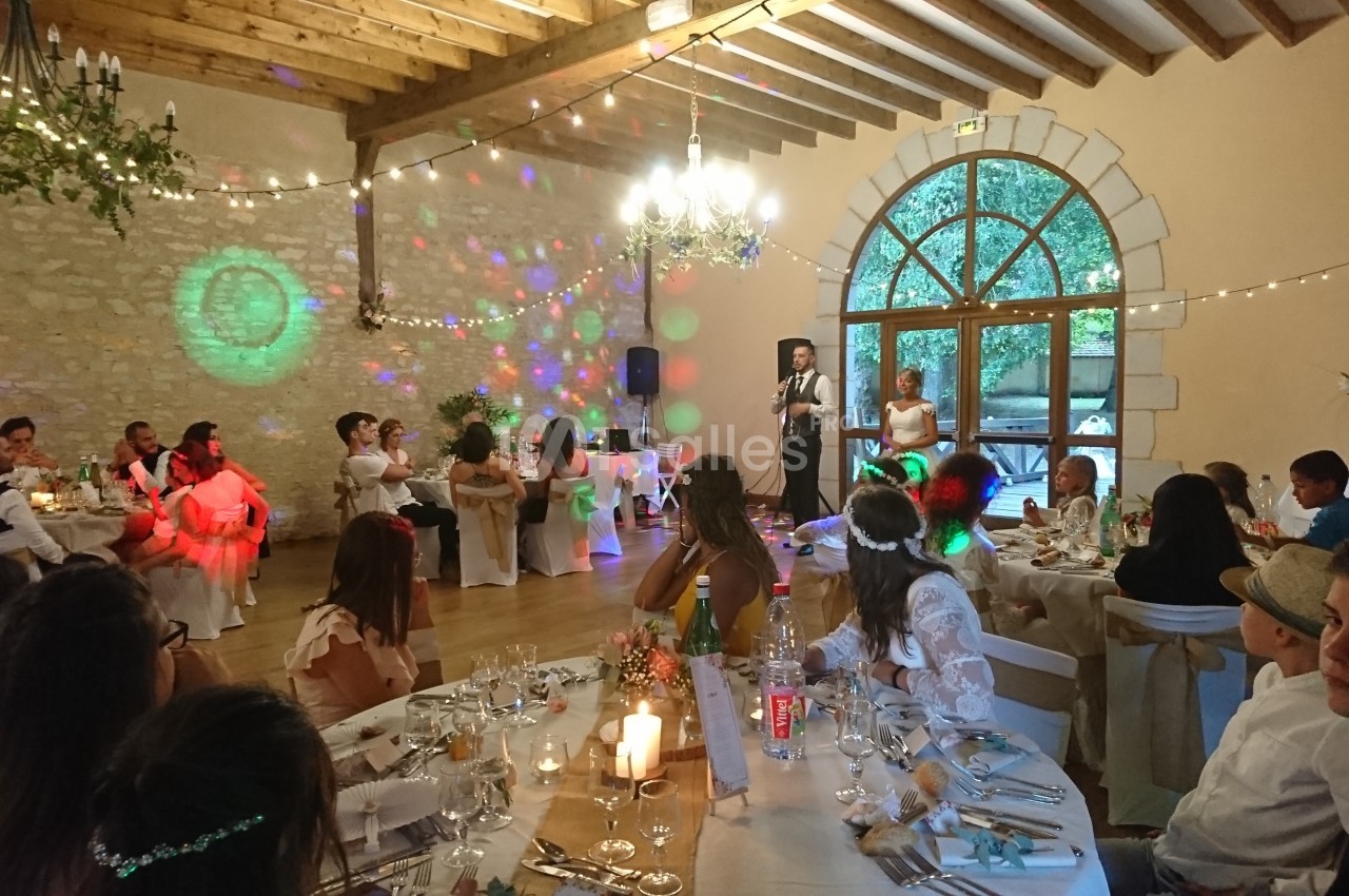 Salle de réception décorée pour un mariage, avec invités assis à table et un couple faisant un discours.