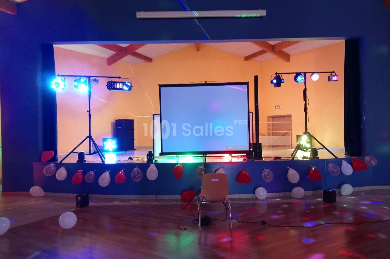 Scène décorée avec ballons, éclairages colorés, écran de projection et projecteurs dans une salle polyvalente.