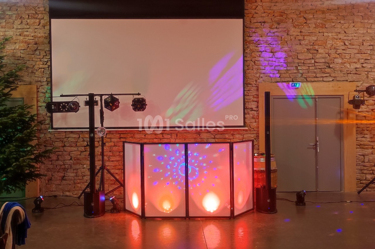 Salle décorée pour un événement, avec mur en pierre, éclairages colorés, écran blanc et sapin dans un coin.