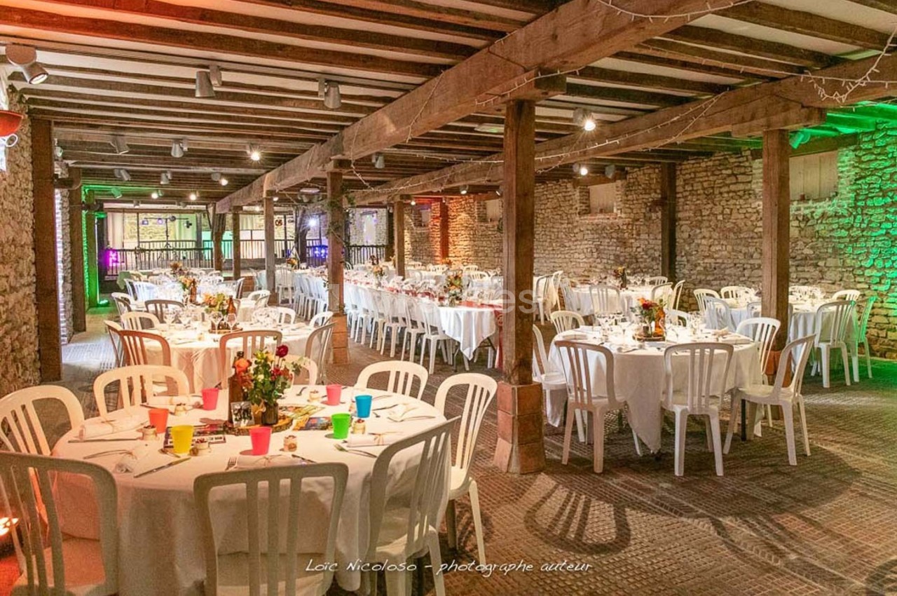 Salle de réception rustique avec tables rondes dressées, chaises blanches et décorations florales colorées.