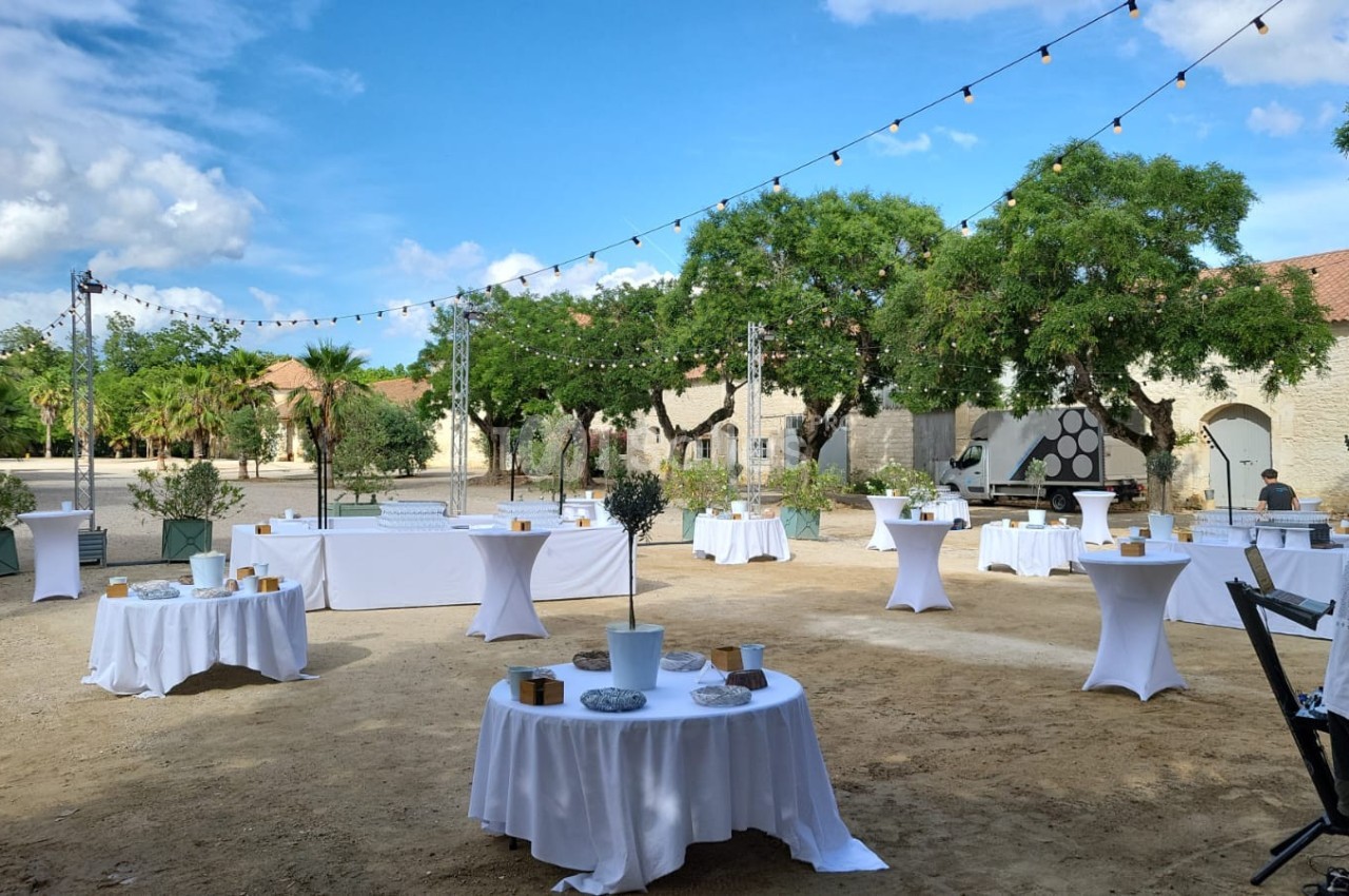 Tables rondes dressées en extérieur sous des guirlandes lumineuses, entourées d'arbres et de bâtiments en pierre.