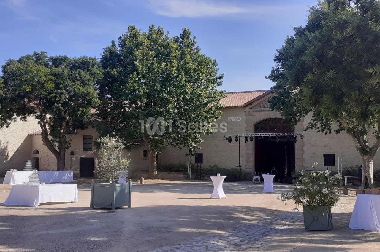 Cour extérieure avec tables hautes et nappes blanches, entourée d'arbres et de bâtiments en pierre sous un ciel dégagé.
