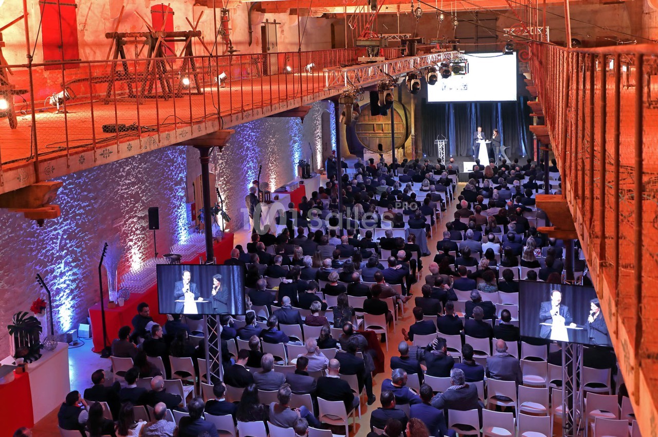 Salle comble avec des participants assis, regardant une présentation sur scène, éclairée par des projecteurs.