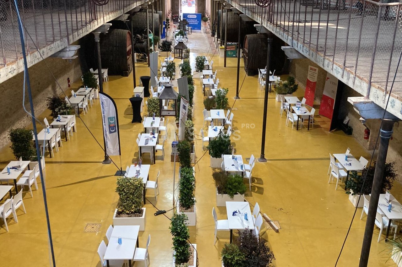 Salle avec des tables et chaises blanches alignées, entourées de plantes, sous une structure métallique ouverte.