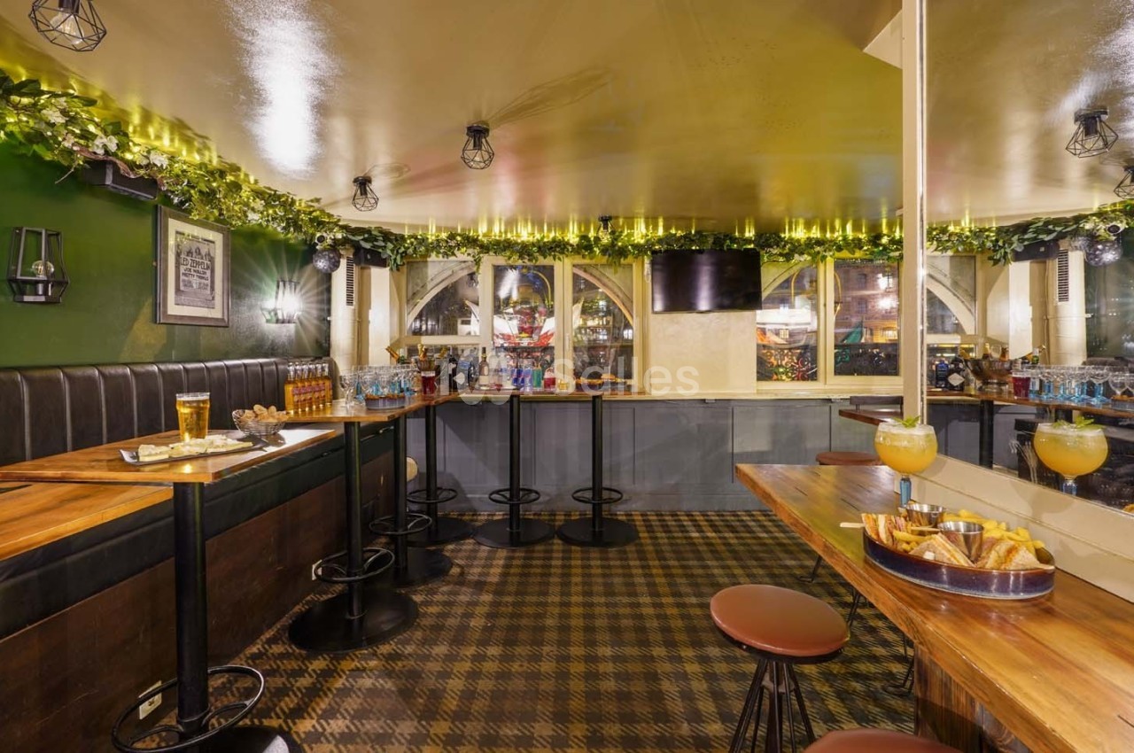 Salle de bar chaleureuse avec tables en bois, tabourets, guirlandes lumineuses et boissons disposées sur le comptoir.