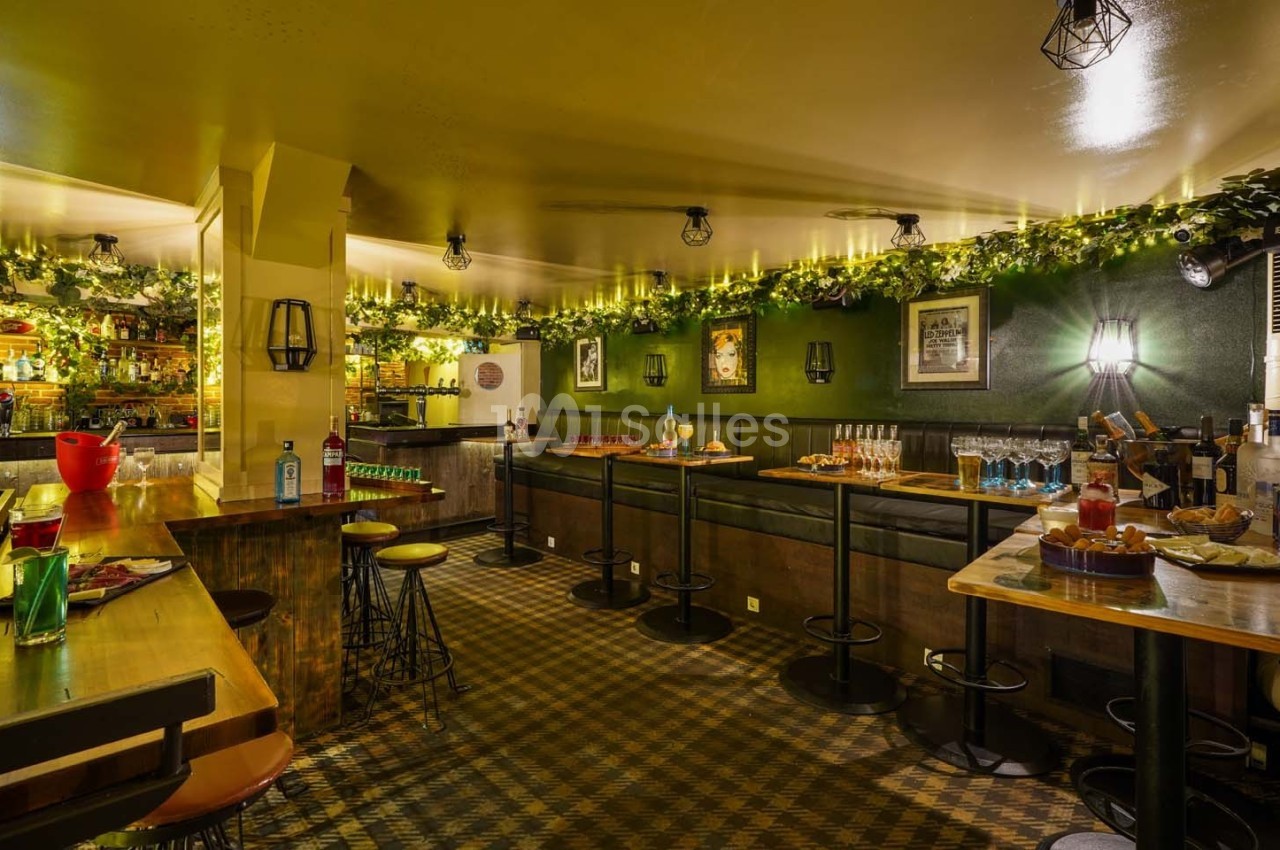 Salle de bar chaleureuse avec tables hautes, tabourets, décorations végétales et lumières tamisées.