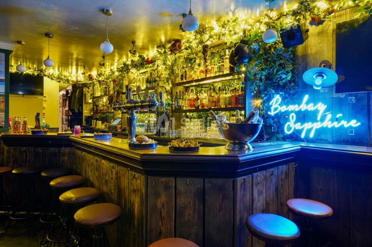 Bar en bois avec tabourets, éclairage chaleureux, étagères remplies de bouteilles et enseigne lumineuse bleue.
