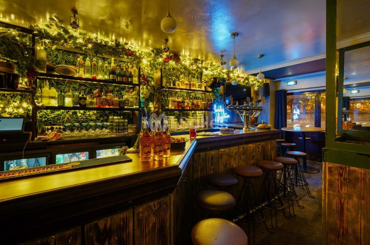 Intérieur d'un bar chaleureux avec comptoir en bois, tabourets hauts, étagères garnies de bouteilles et éclairage tamisé.