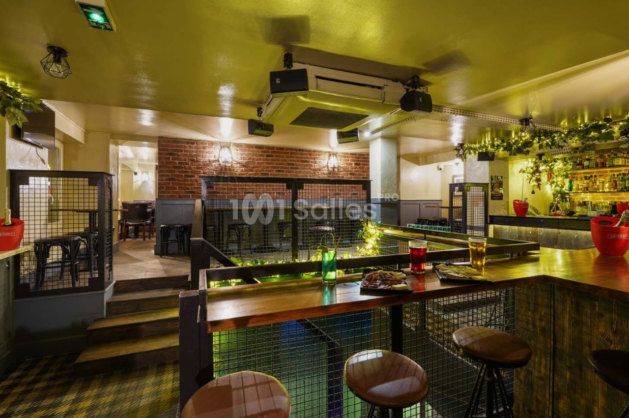 Intérieur d'un bar avec un comptoir en bois, des tabourets, un éclairage chaleureux et une décoration végétale.