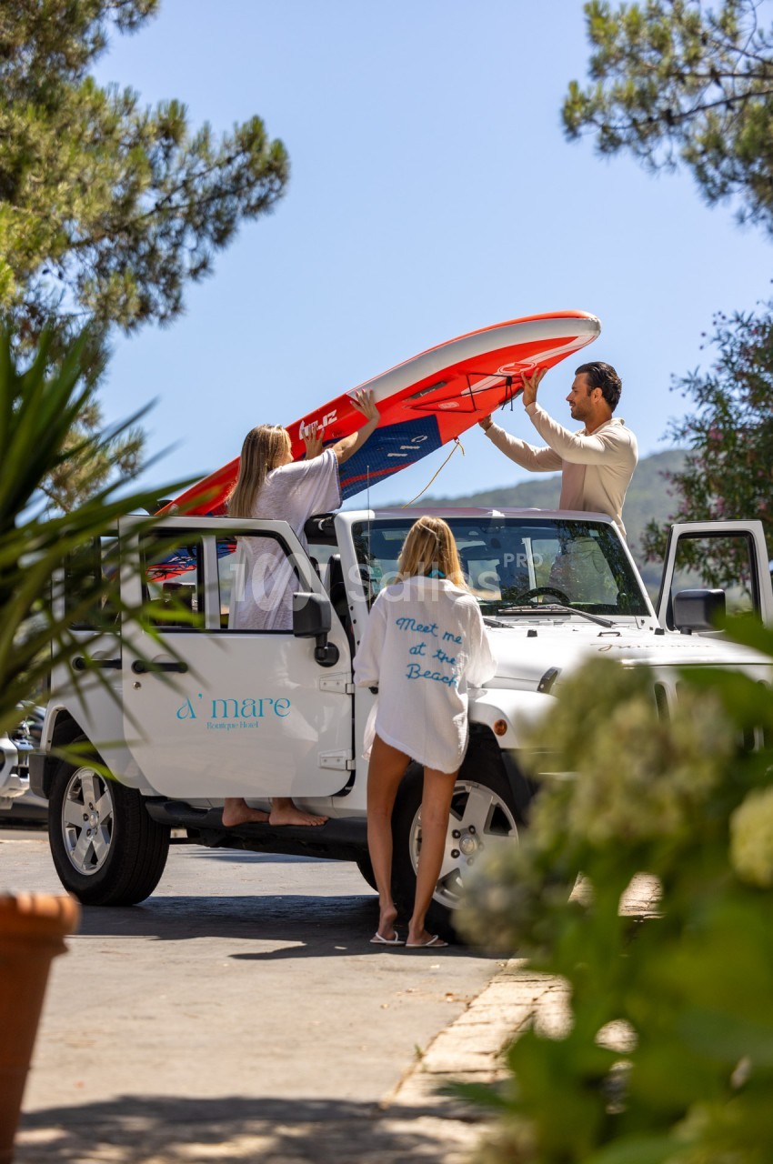 Deux femmes et un homme chargent une planche de surf rouge sur un 4x4 blanc, entourés de végétation sous un ciel bleu.