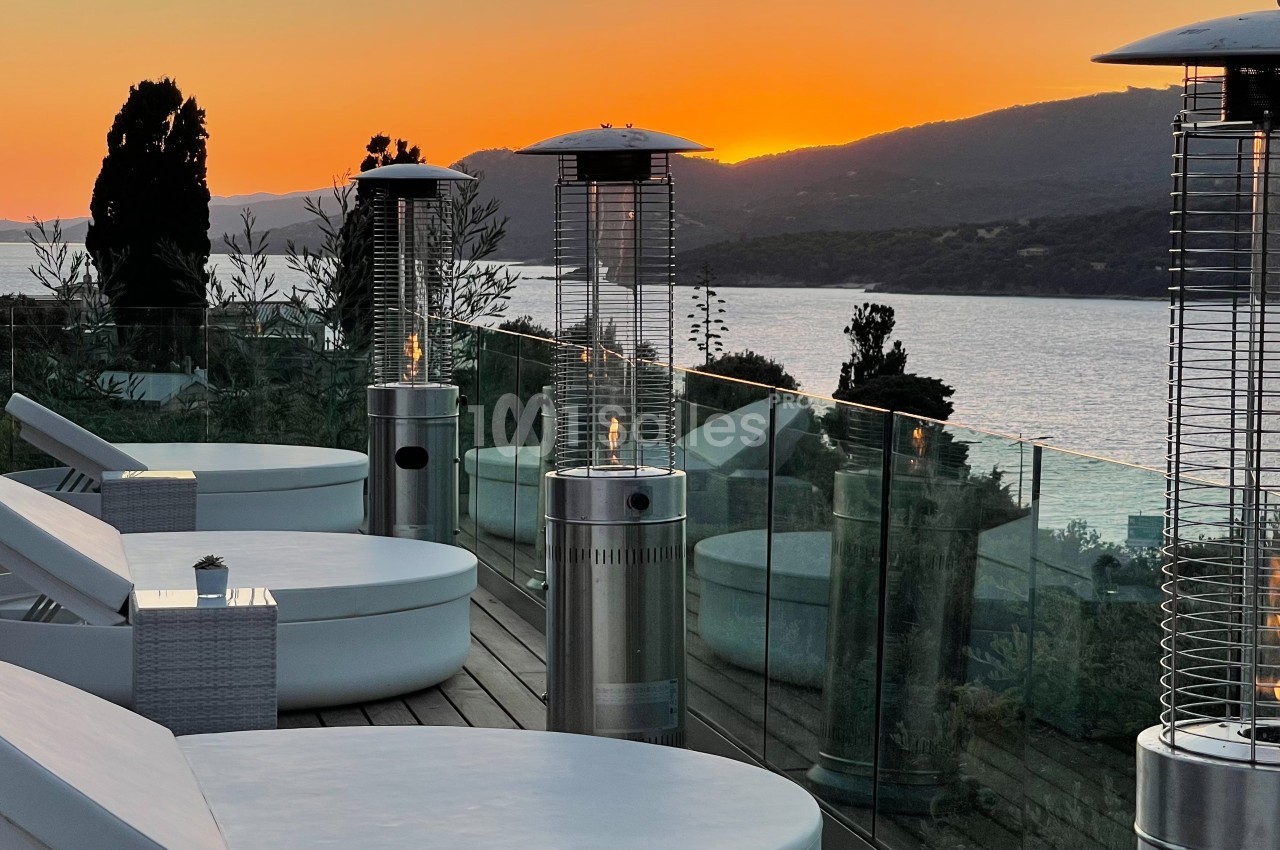 Terrasse avec chaises longues, chauffages extérieurs et vue sur un lac entouré de collines au coucher du soleil.