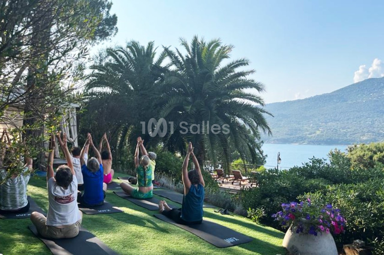 Personnes pratiquant le yoga en plein air sur une pelouse, entourées de végétation avec vue sur la mer et les montagnes.