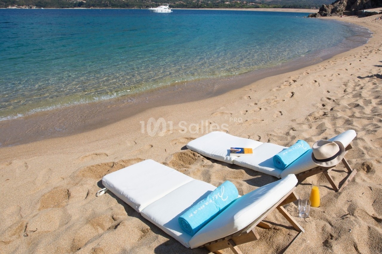 Deux chaises longues avec serviettes bleues sur une plage de sable, face à une mer calme et des collines en arrière-plan.