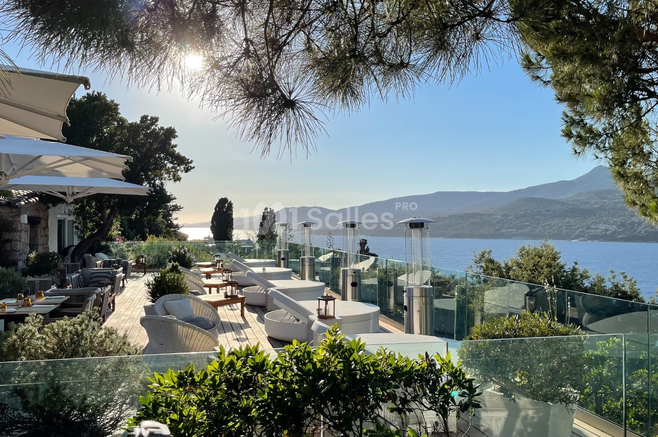 Terrasse ensoleillée avec mobilier élégant, vue sur la mer et collines boisées à l'arrière-plan.