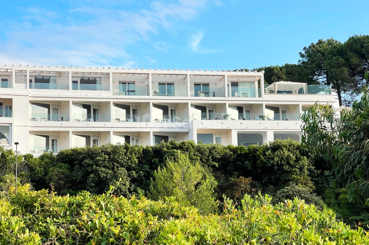 Bâtiment moderne blanc à plusieurs étages avec balcons, entouré de végétation dense sous un ciel bleu.