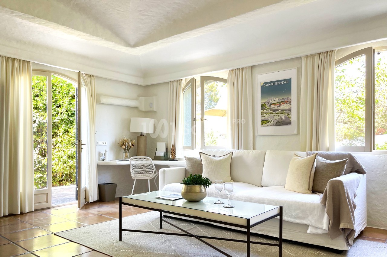Salon lumineux avec canapé blanc, table basse, bureau près de la fenêtre et vue sur un jardin verdoyant.