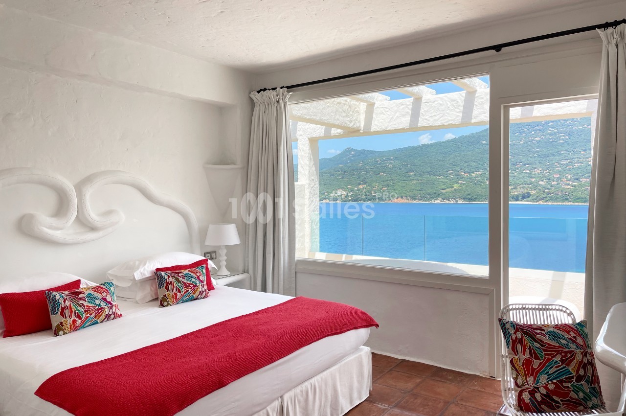 Chambre lumineuse avec lit double, décor blanc et rouge, vue sur la mer et montagnes à travers une grande baie vitrée.