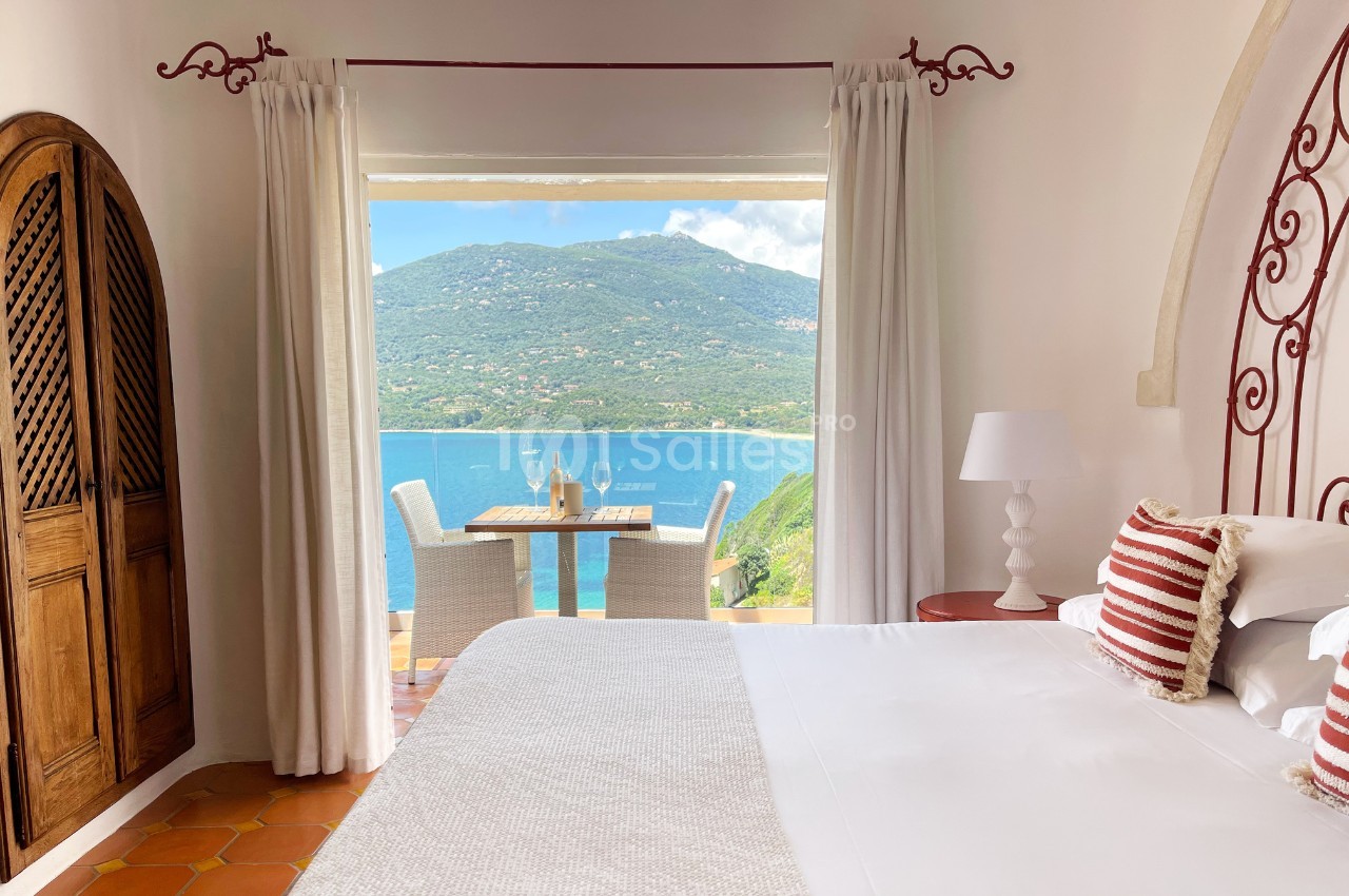 Chambre lumineuse avec lit, rideaux ouverts sur une terrasse offrant une vue sur la mer et des montagnes verdoyantes.