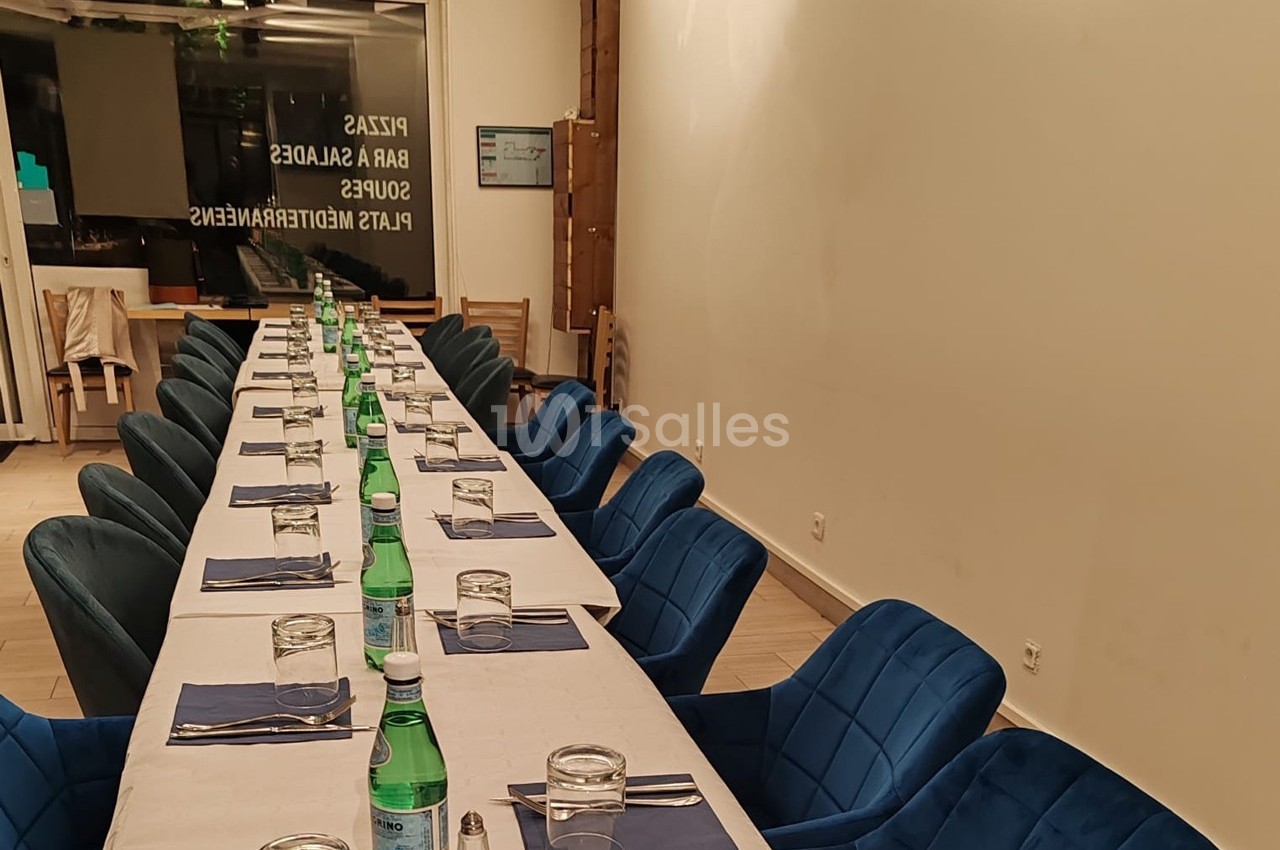Salle de restaurant avec une longue table dressée, nappes blanches, chaises bleues et bouteilles d'eau minérale.