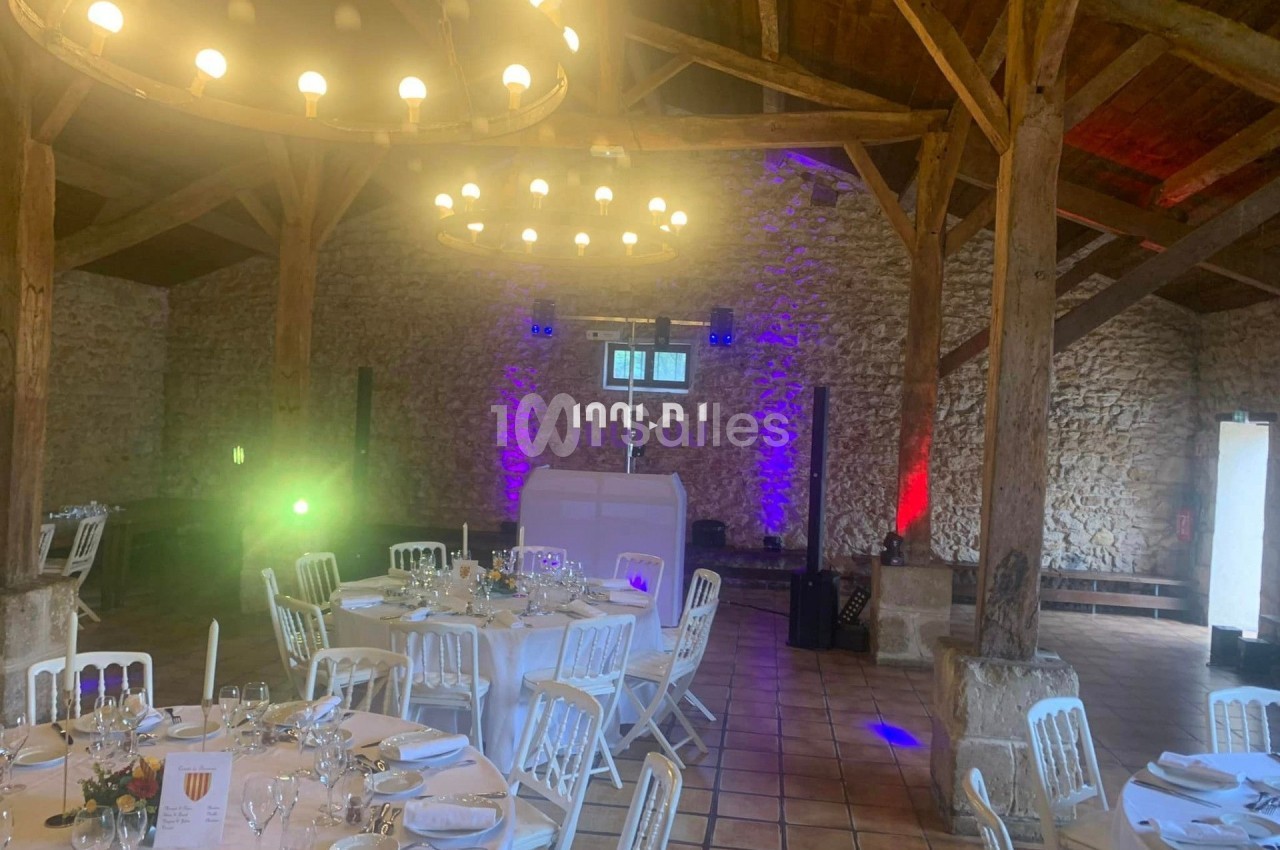 Salle de réception en pierre avec tables dressées, éclairage coloré et poutres apparentes en bois.