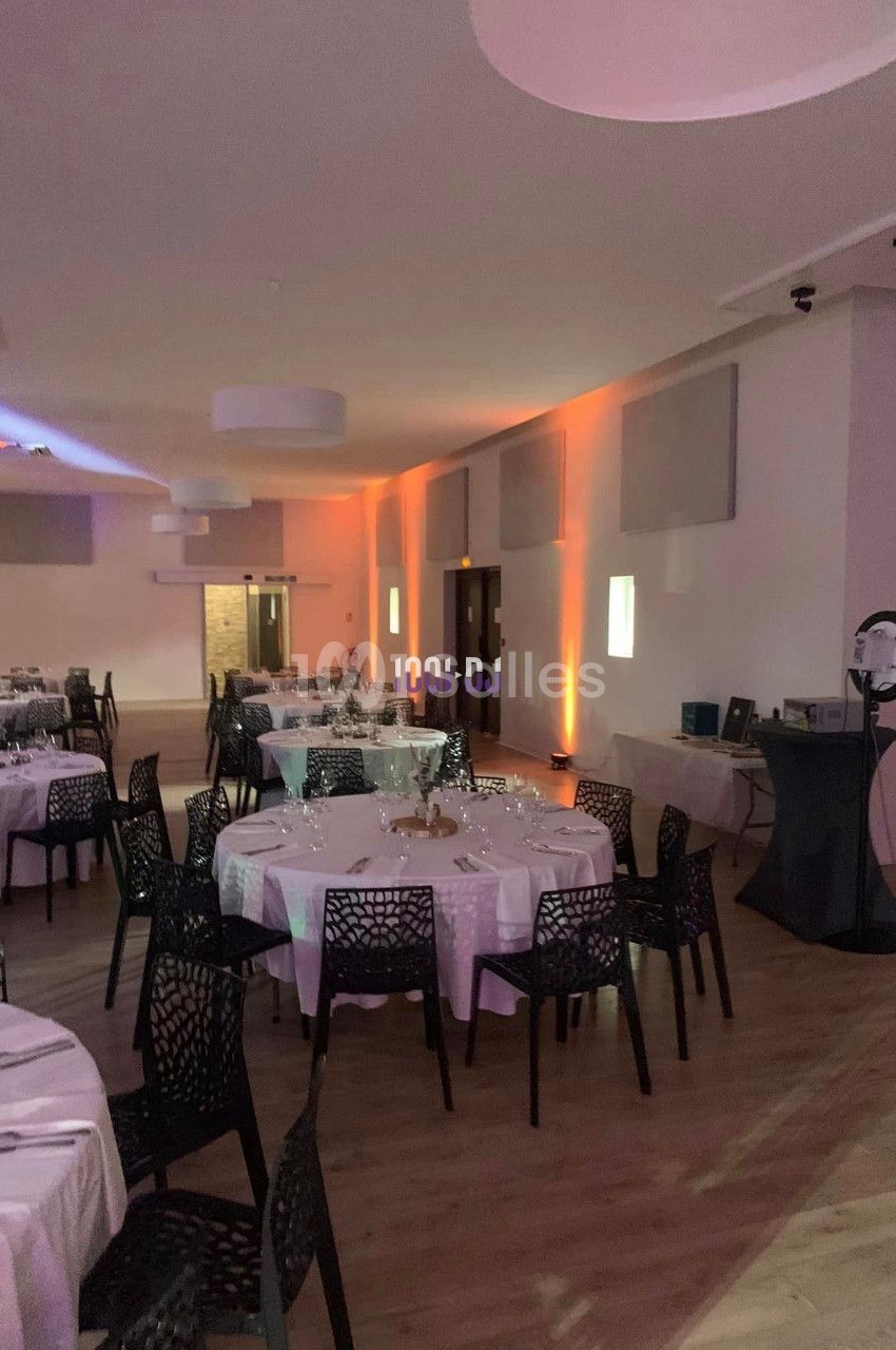 Salle de réception décorée avec des tables rondes dressées, chaises noires et éclairage tamisé.