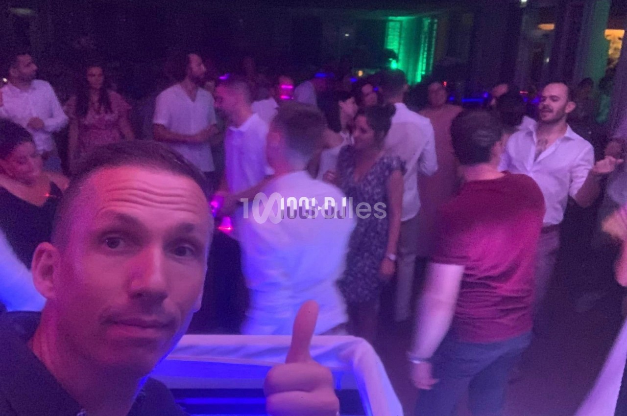 Un homme fait un signe de pouce levé dans une soirée animée avec des personnes dansant sous des lumières colorées.