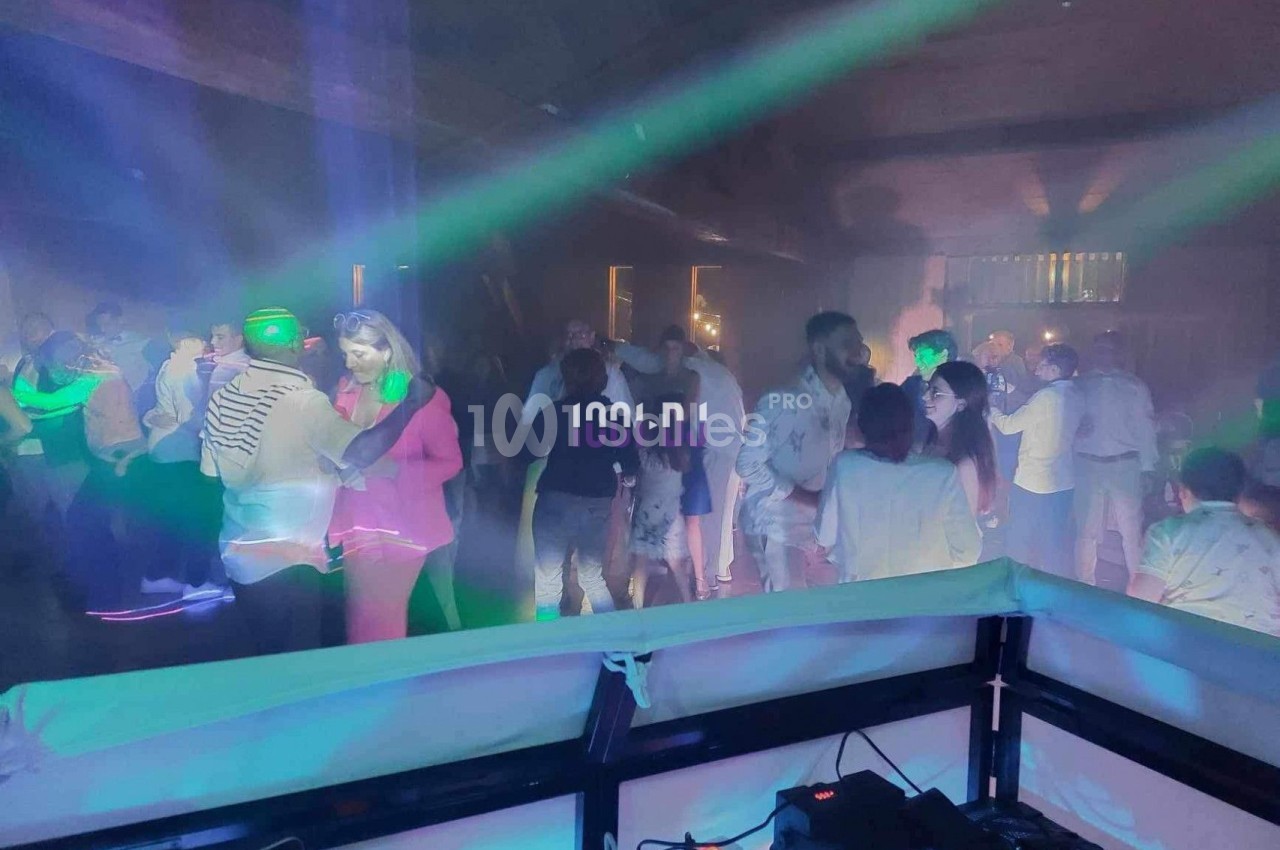 Des personnes dansent sous des lumières colorées dans une ambiance festive et enfumée, vue depuis une table de DJ.