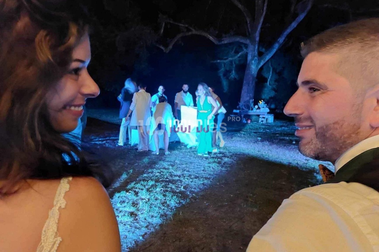 Un couple souriant s'observe lors d'une soirée en plein air, avec des invités discutant sous un éclairage nocturne.