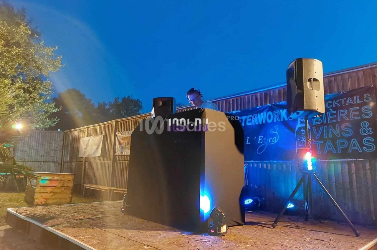 Un DJ mixe sur une scène extérieure éclairée en soirée, entourée de panneaux et d'enceintes.