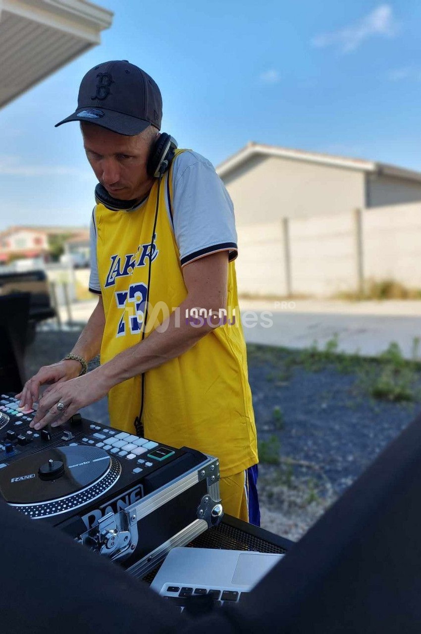 Un DJ mixant en extérieur, portant un maillot jaune et un casque audio, avec des maisons en arrière-plan.