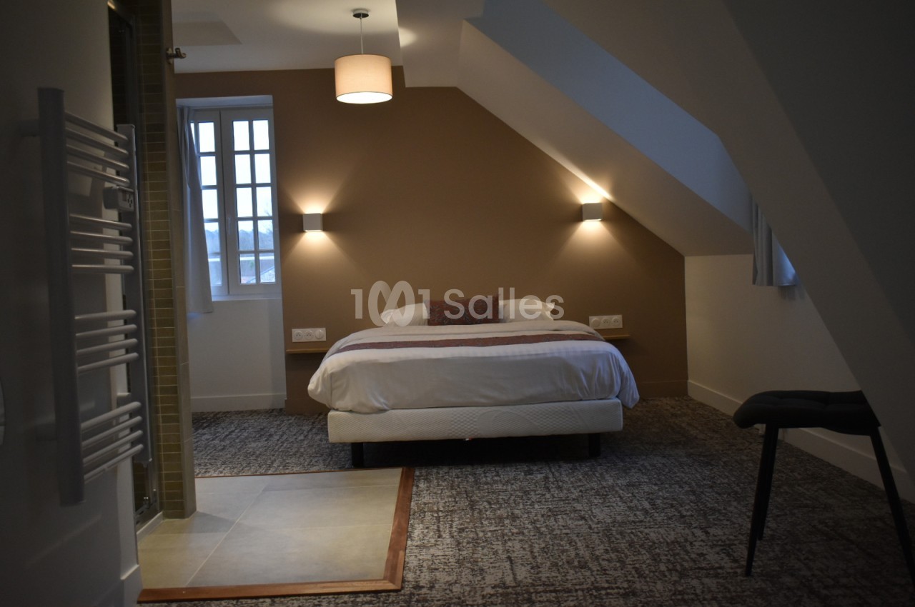 Chambre mansardée avec un lit double, éclairage mural, moquette au sol et salle de bain attenante visible à gauche.
