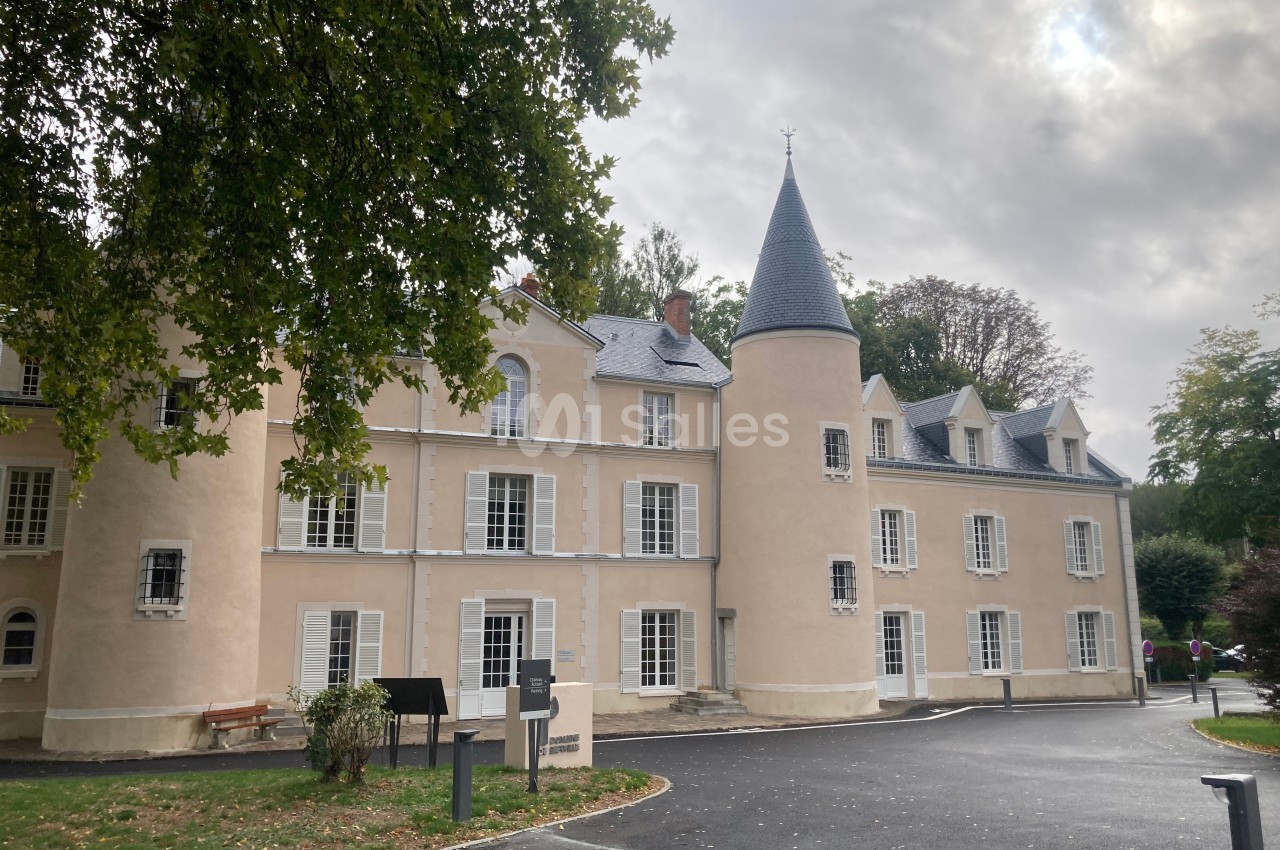 Façade d'un bâtiment historique beige avec une tour ronde et des fenêtres blanches, entouré d'arbres et d'une allée.