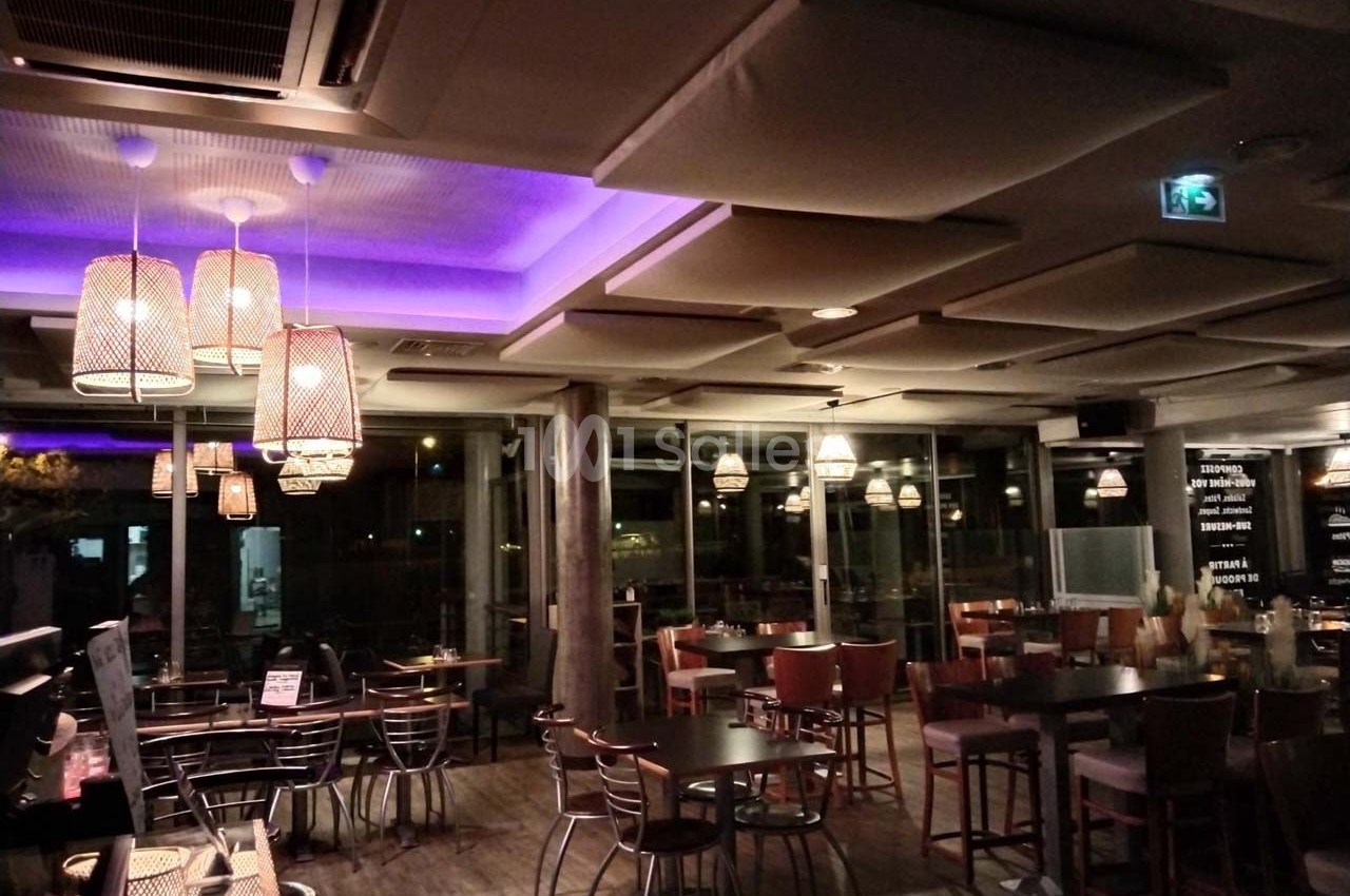 Salle de restaurant moderne avec éclairage tamisé, plafonniers suspendus et grandes baies vitrées donnant sur l'extérieur.