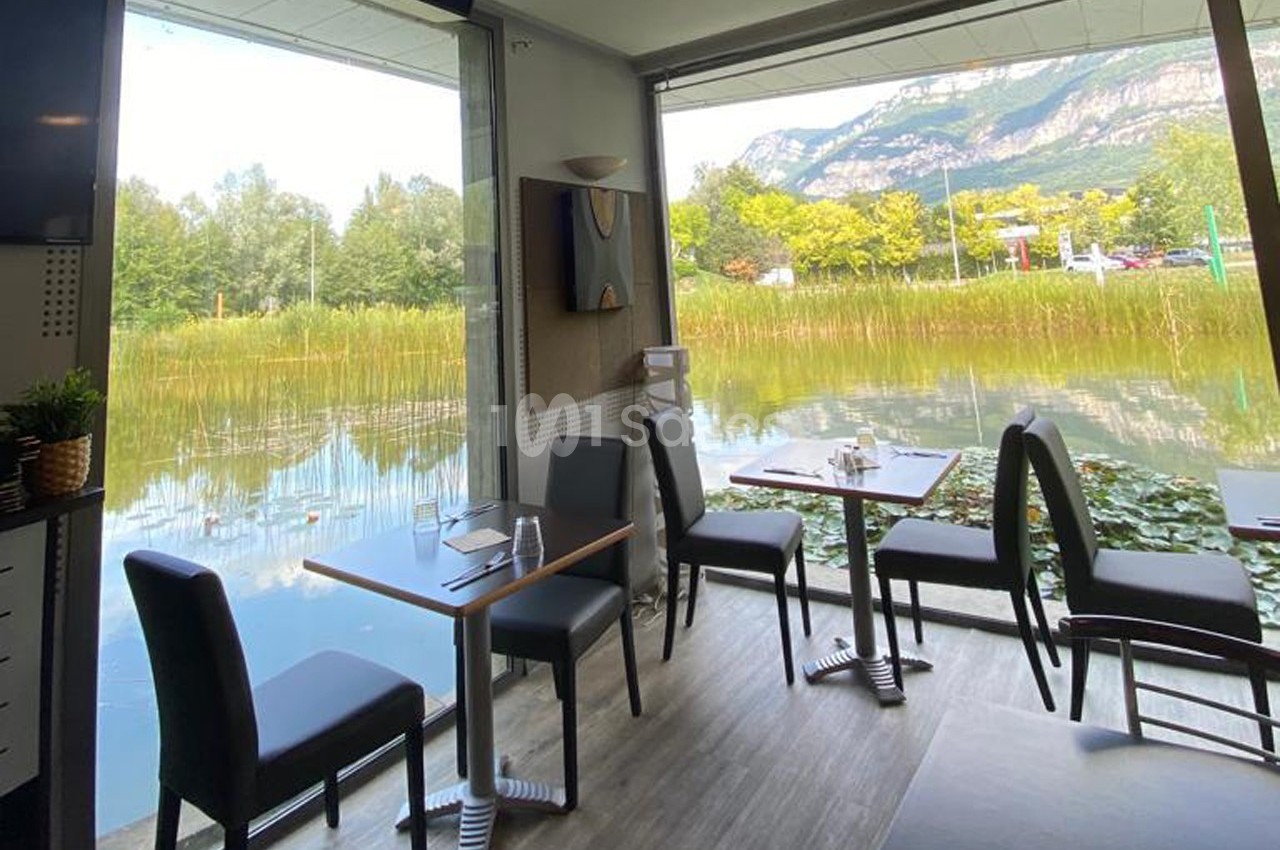 Salle de restaurant avec baies vitrées donnant sur un étang bordé de végétation et des montagnes en arrière-plan.