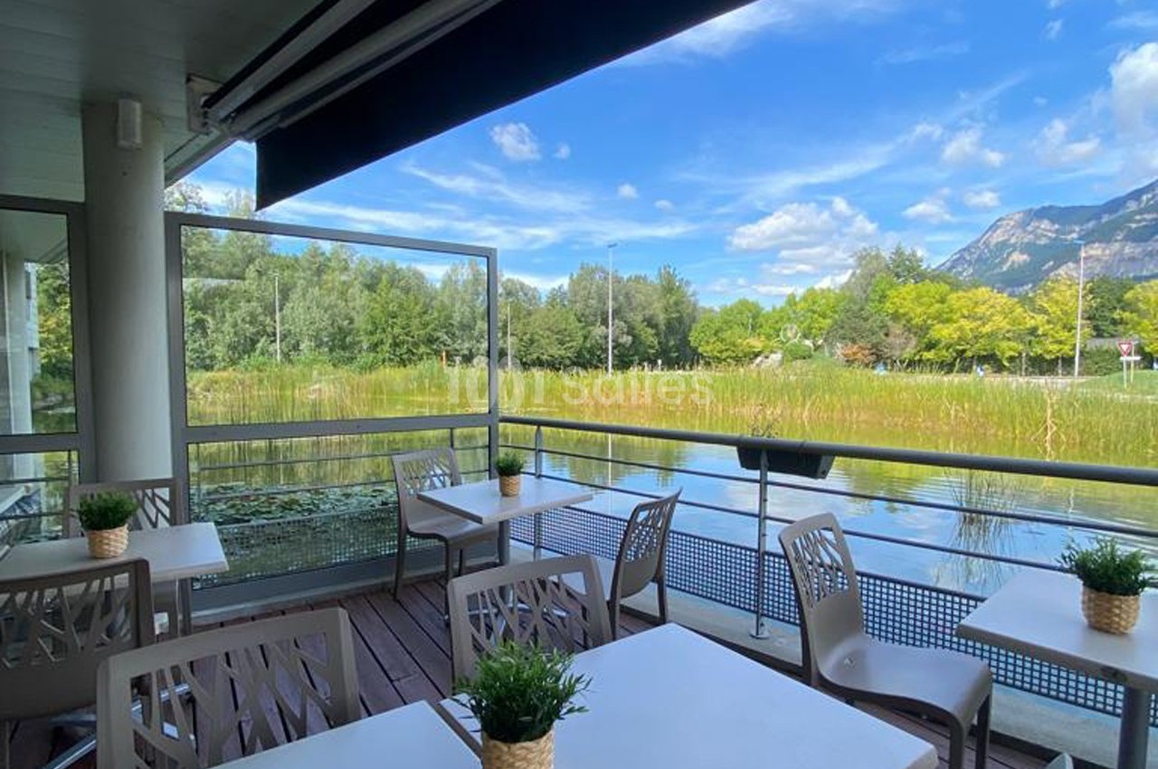 Terrasse avec tables et chaises donnant sur un étang bordé de végétation, sous un ciel bleu partiellement nuageux.