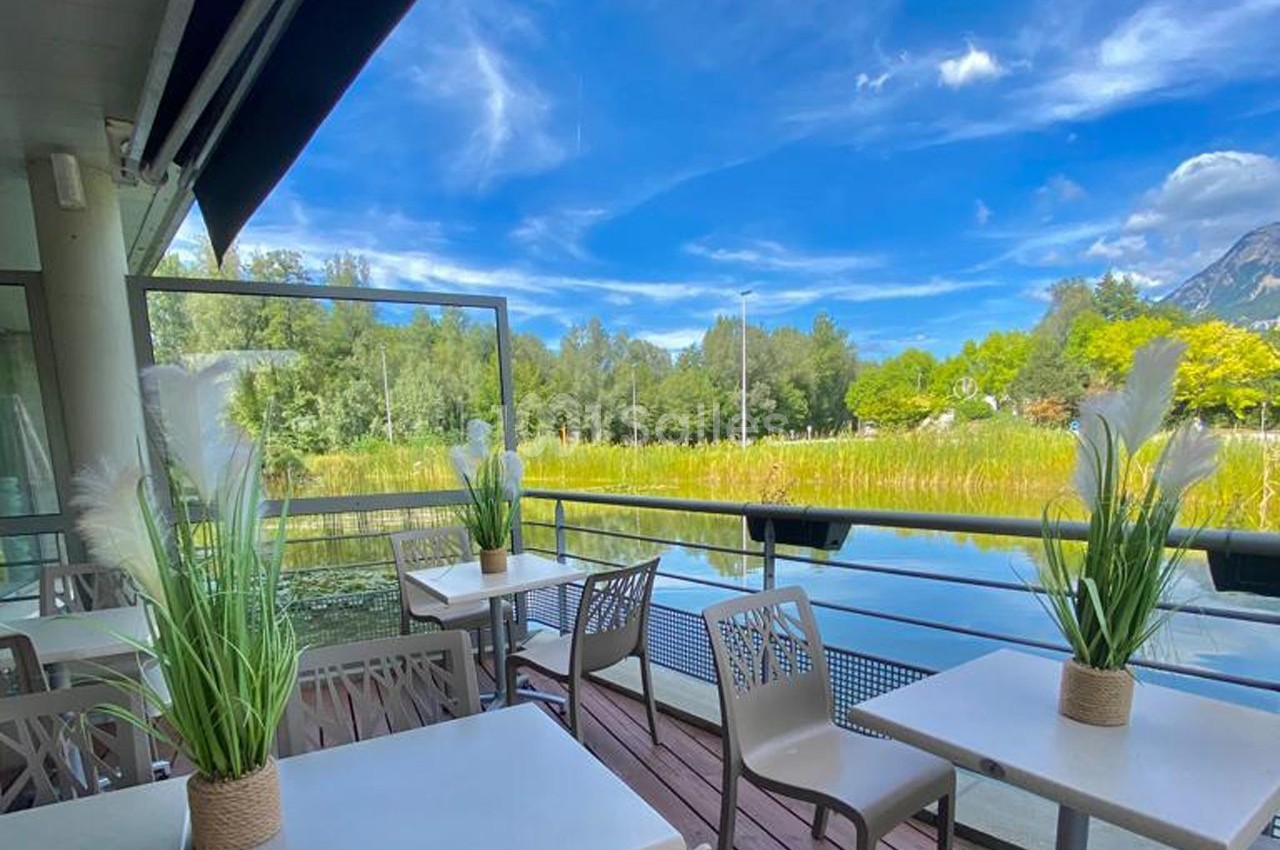 Terrasse avec tables et chaises donnant sur un paysage naturel avec un lac, des arbres et des montagnes sous un ciel bleu.