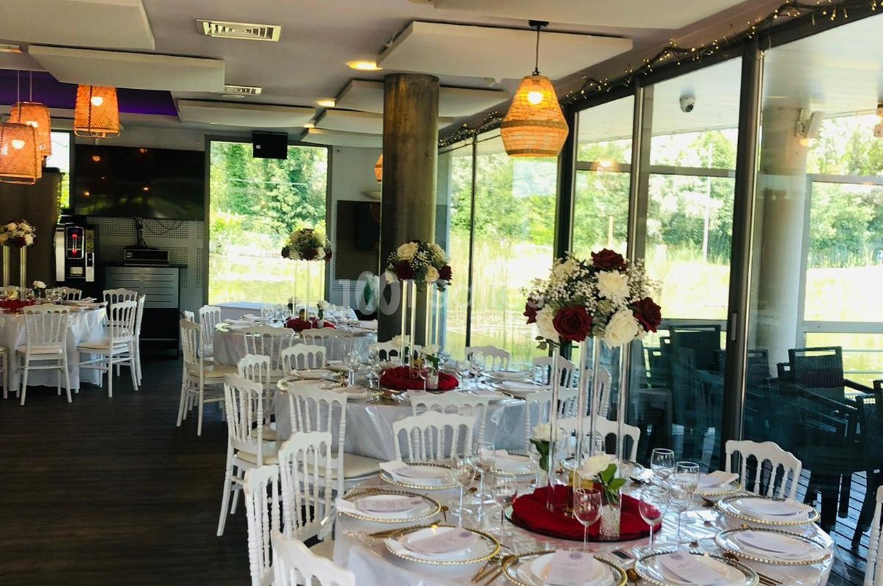 Salle de réception lumineuse avec tables rondes décorées de fleurs rouges et blanches, vue sur un extérieur verdoyant.