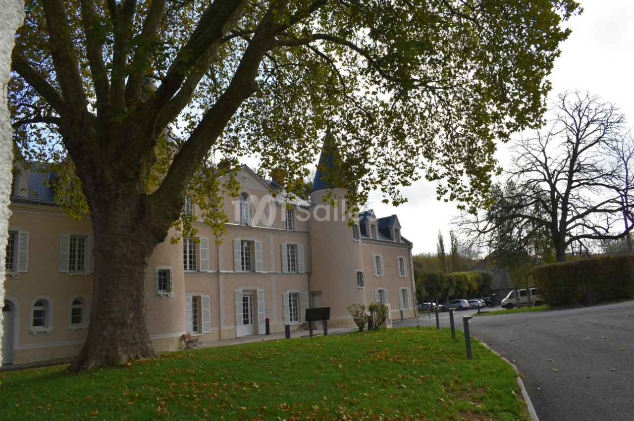 Façade d'un bâtiment historique beige avec tours, entouré d'arbres et d'une pelouse, sous un ciel partiellement couvert.