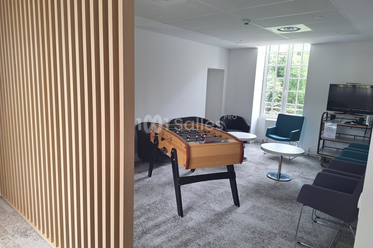 Salle de détente avec baby-foot, fauteuils, tables basses et télévision, éclairée par une grande fenêtre.