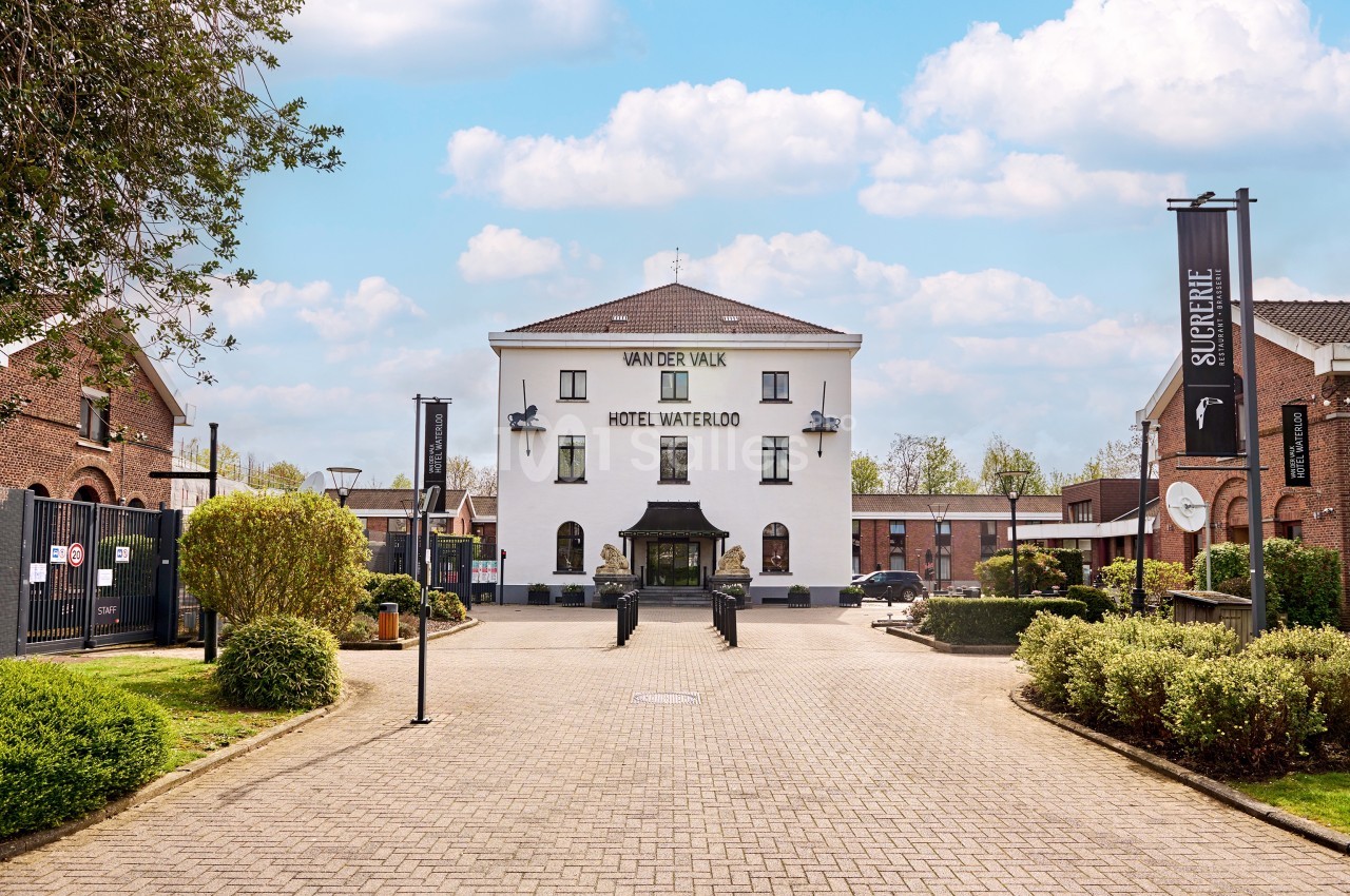 Façade de l'hôtel Van der Valk Waterloo, entourée de verdure et de bâtiments en briques sous un ciel bleu.