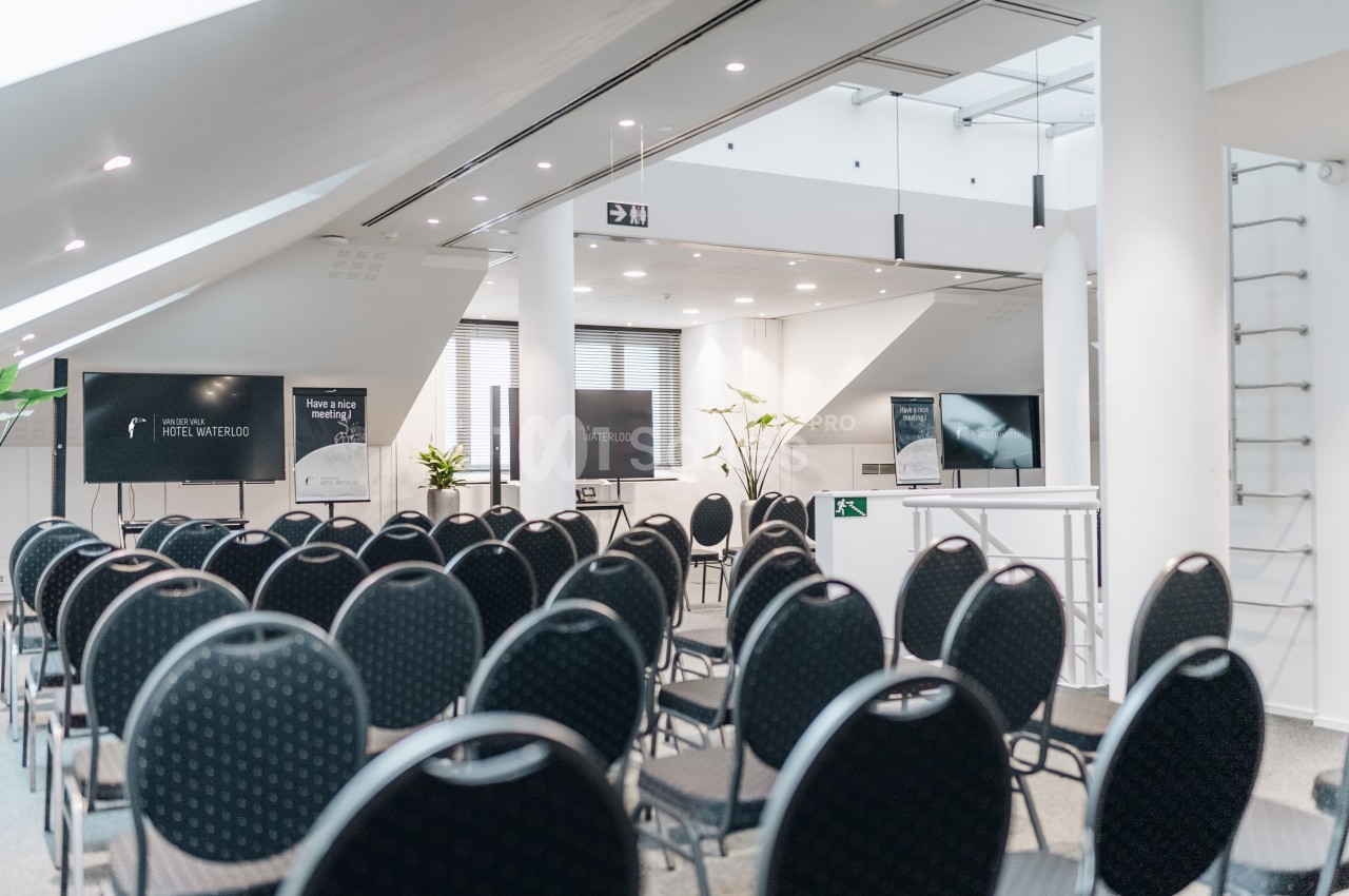 Salle de conférence moderne avec des rangées de chaises noires vides, écrans et éclairage naturel.