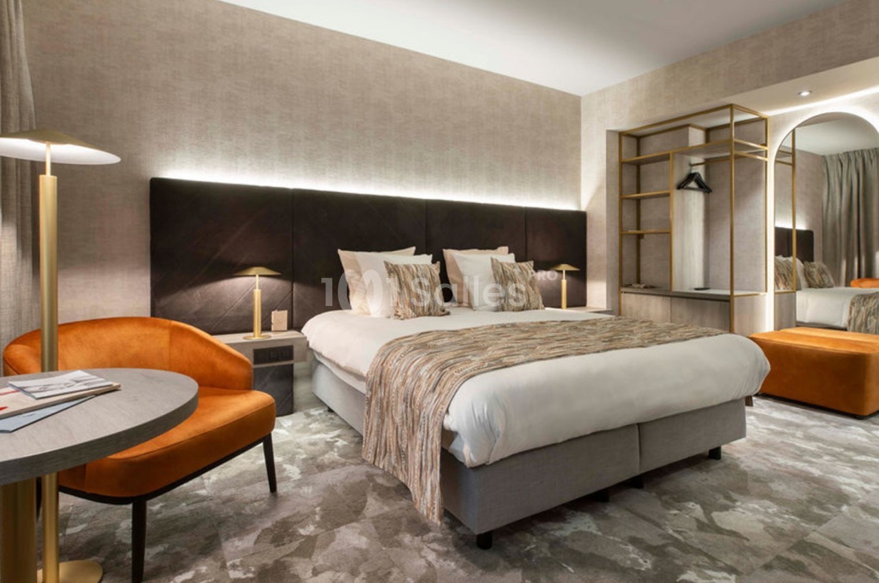 Chambre moderne avec lit double, fauteuil orange, bureau, éclairage doux et miroir près d'une armoire ouverte.