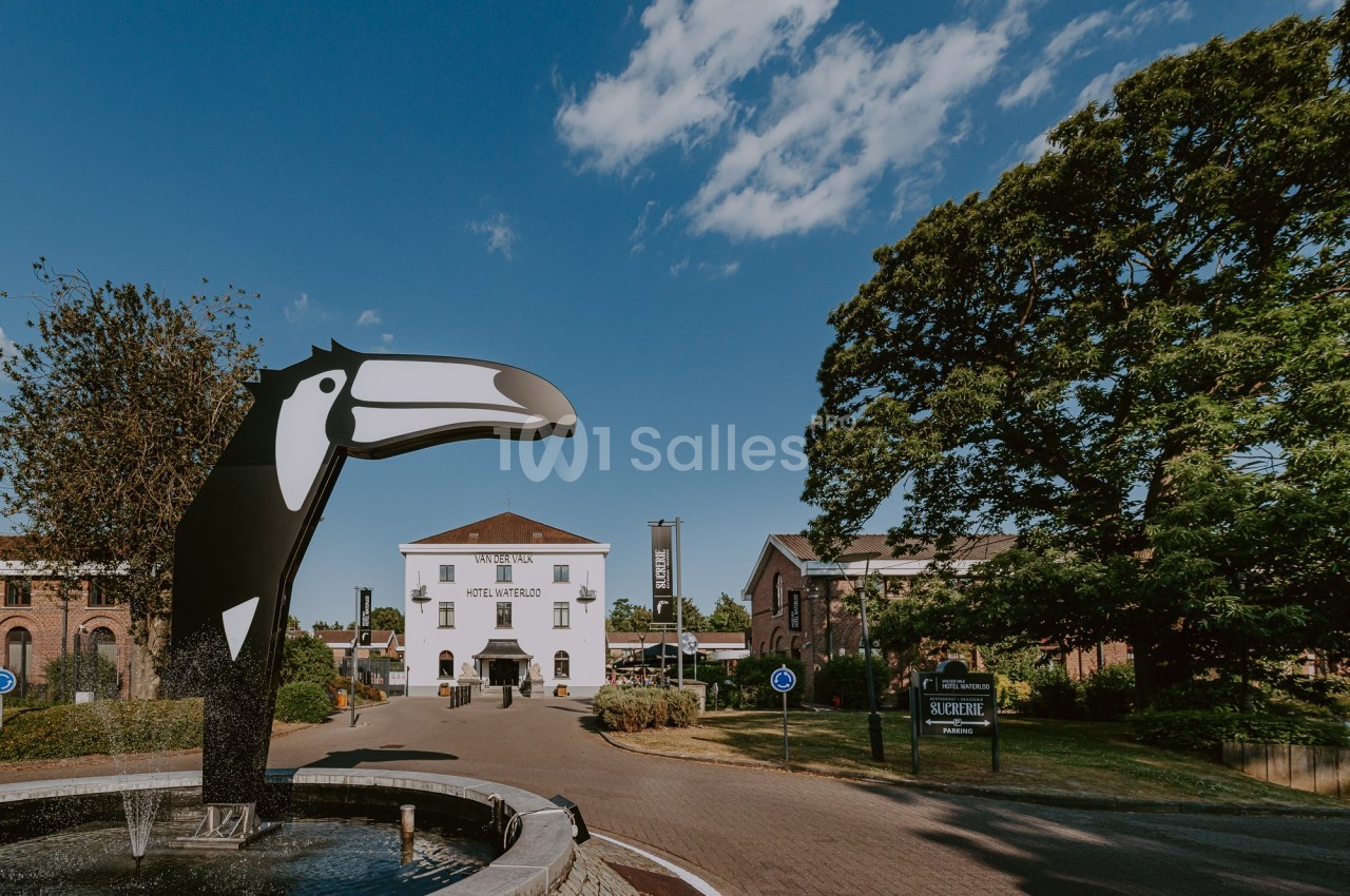Statue d'un toucan près d'une fontaine, avec des bâtiments en briques et un ciel dégagé en arrière-plan.