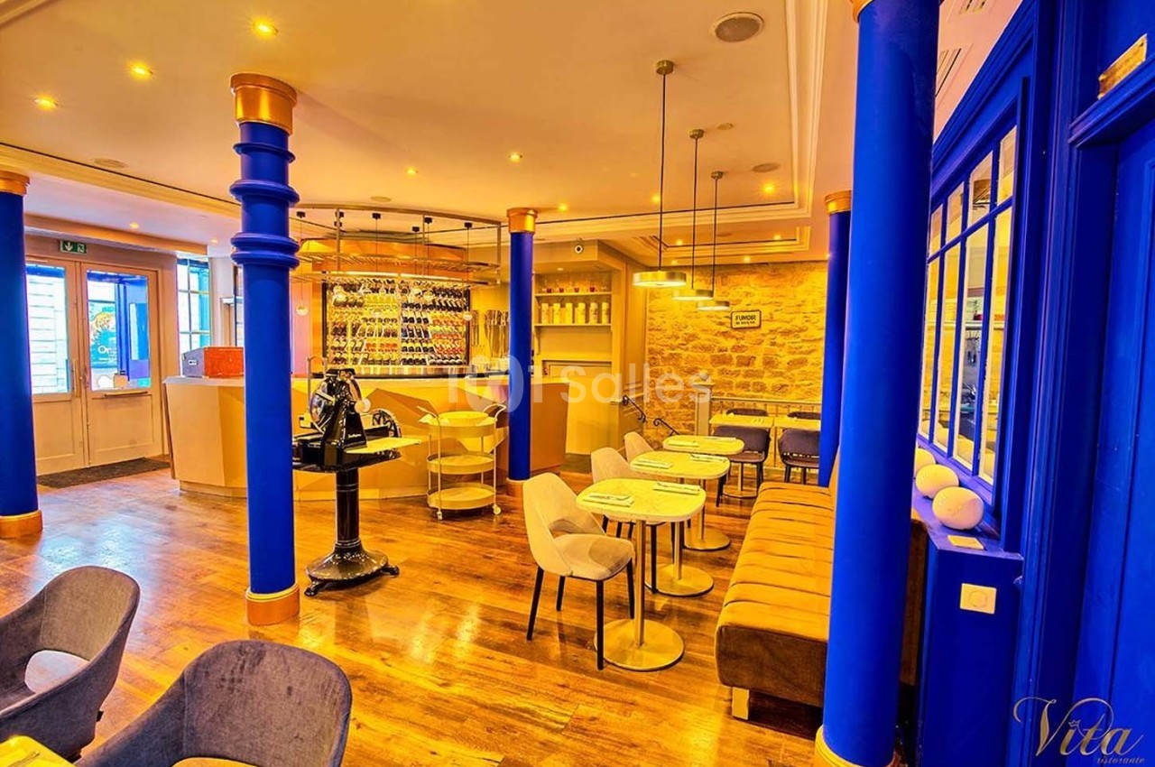 Salle de restaurant lumineuse avec murs en pierre, colonnes bleues, mobilier moderne et bar à vin en arrière-plan.