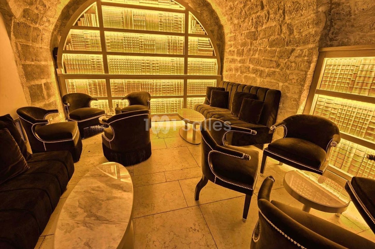 Salon voûté en pierre avec fauteuils en velours noir, tables basses en marbre et étagères lumineuses en arrière-plan.