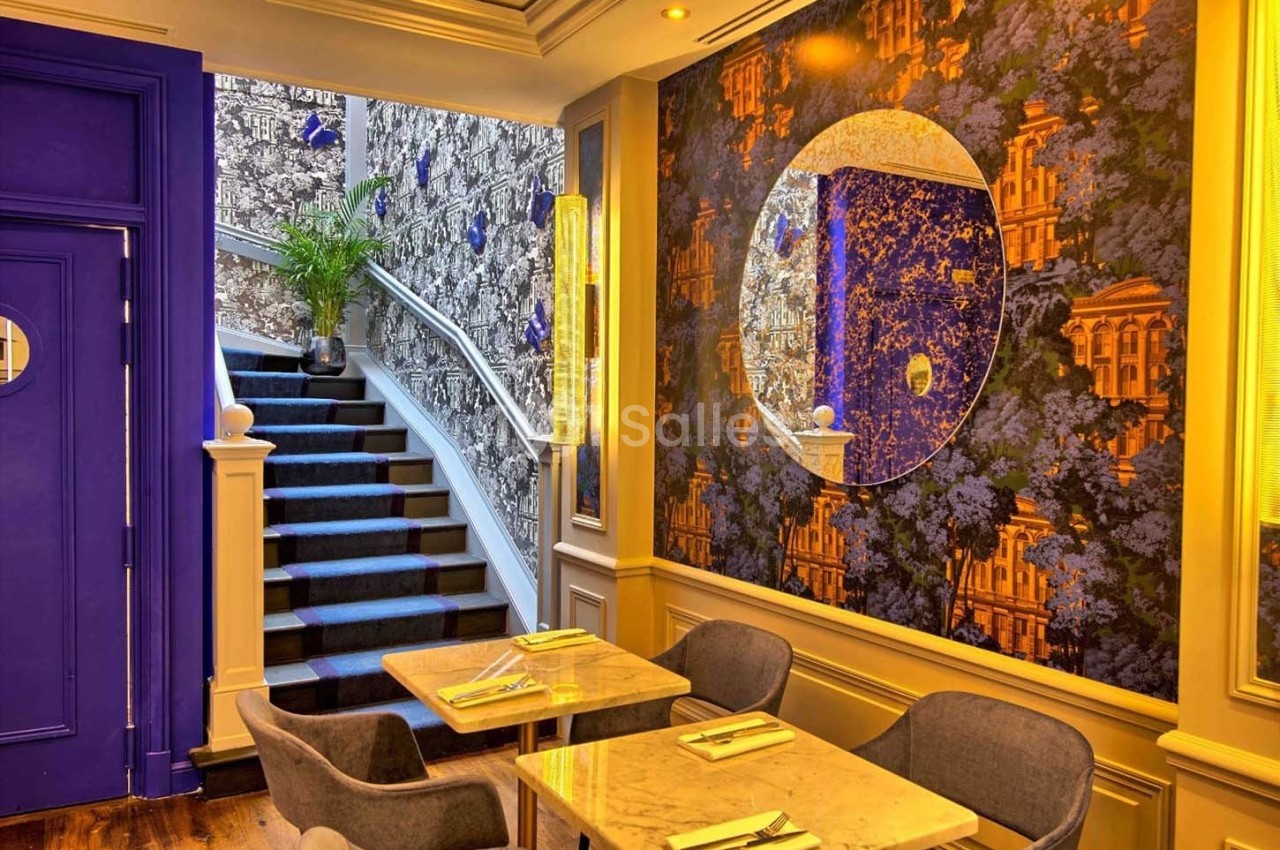 Salle de restaurant colorée avec tables dressées, escalier tapissé menant à l'étage et miroir décoratif sur un mur.