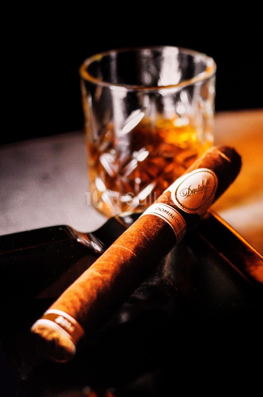 Un cigare posé sur un cendrier noir avec un verre de whisky ambré en arrière-plan.