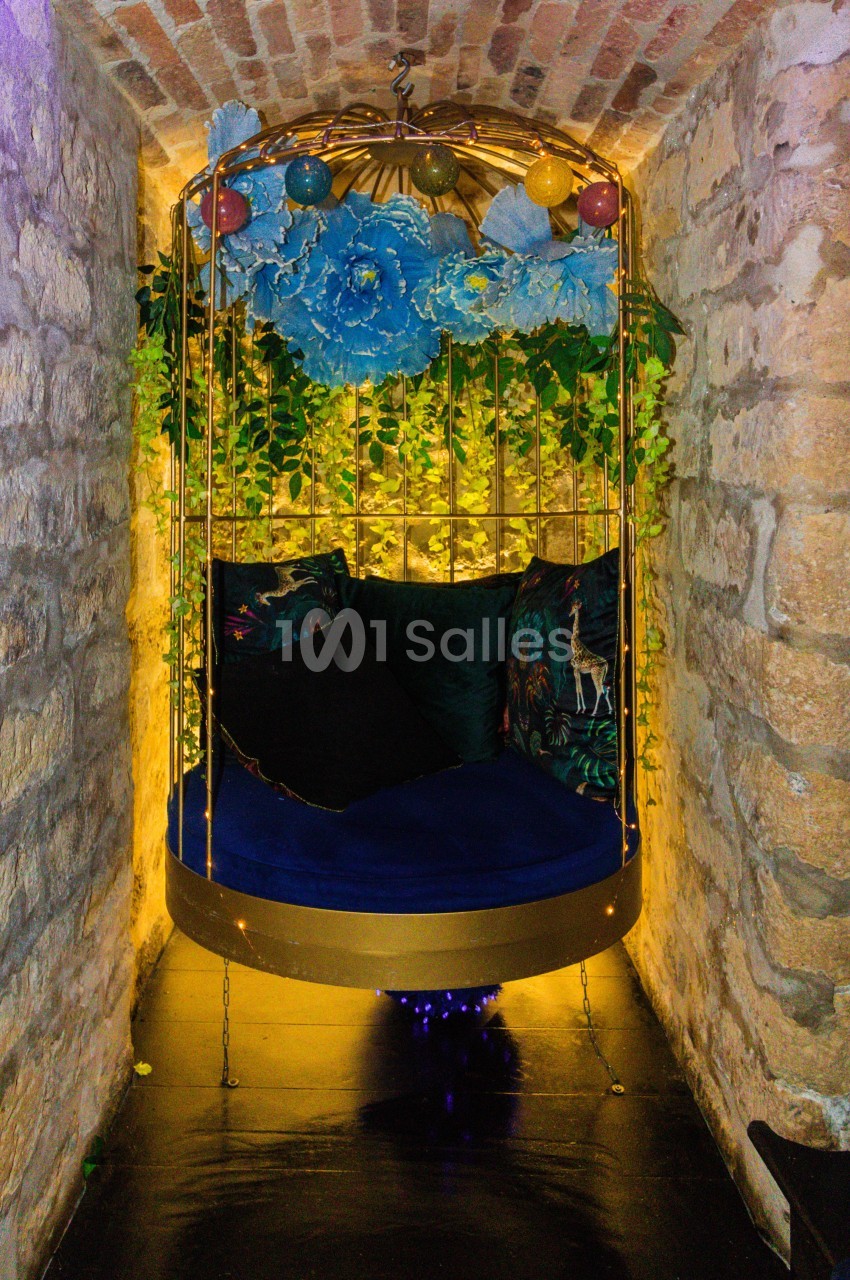Balancelle dorée suspendue, entourée de fleurs, feuillages et éclairages chaleureux, dans une alcôve en pierre.
